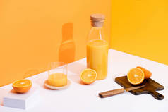 新鲜橙汁在玻璃瓶中，靠近成熟的橙子，木制切菜板，白色表面上有刀片，橙色背景