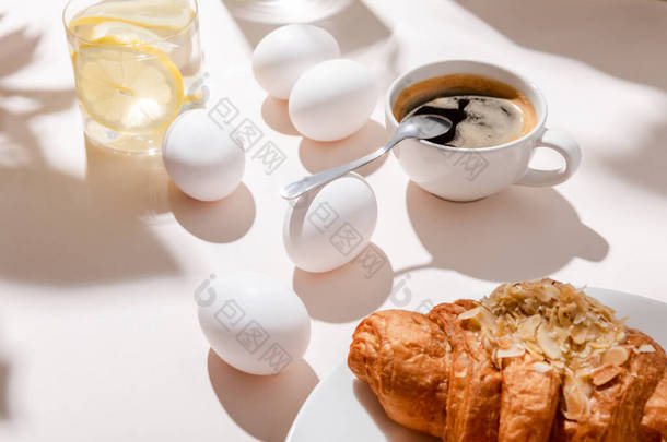鸡蛋、羊角面包、一杯咖啡和一杯柠檬水，早餐放在灰色桌子上，有阴影