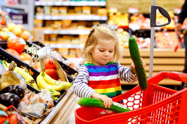 可爱的结结巴巴的姑娘在超级市场推着购物车。小孩在买水果。儿童食品杂货店购物。可爱的婴孩与推车选择新鲜蔬菜在当地商店.