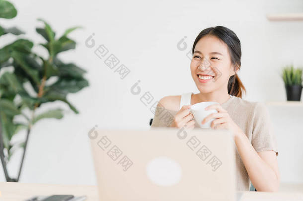 美丽的年轻微笑的亚洲妇女工作在笔记本电脑和喝咖啡在客厅里的家。亚洲商业妇女工作文件财务和计算器在她的家庭办公室。在家享受时光.