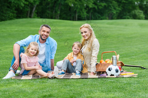 愉快<strong>的</strong>家庭与二个孩子微笑在照相机, 而坐在一起在格子在公园野餐 