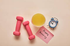 在米色背景上，有快乐的早晨字母、粉红色哑铃、橙汁和小闹钟的便笺的顶视图