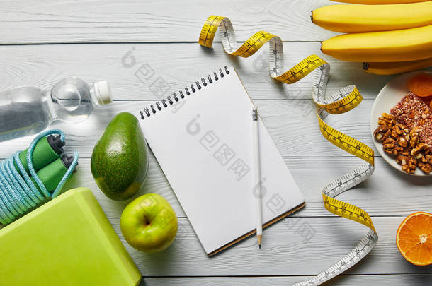 木白色背景上靠近测量带、笔记本和运动器材的减肥食品顶视图