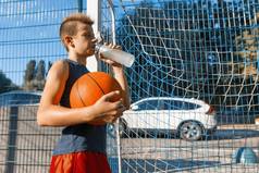 在室外城市篮球场饮用水上与球打街头篮球的少年