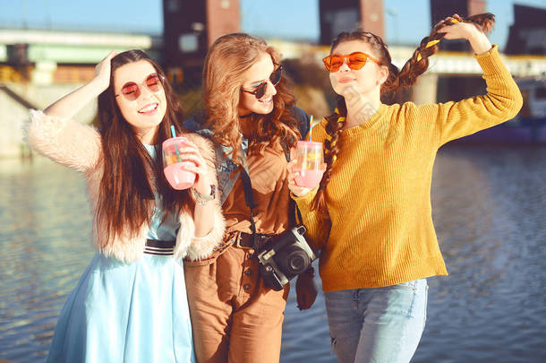 三个女孩在河边对着天空玩得很开心。戴着太阳镜的时尚女孩跳, 高兴, 喝酒, 微笑有乐趣, 疯了。穿着色彩的朋友花夏天的气氛, 时间在一起.