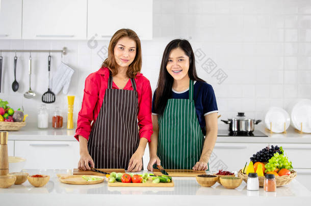 美丽的亚洲妇女在红色衬衫和黑色围裙教她的女儿如何准备蔬菜烹饪在白色清洁现代厨房.