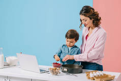 微笑的母亲与小儿子添加红莓烘焙形式, 同时一起做饭在厨房的桌子与烹饪食材和笔记本电脑在双色背景 