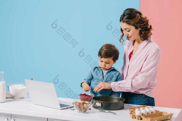 微笑的母亲与小儿子添加红莓烘焙形式, 同时一起做饭在厨房的桌子与烹饪<strong>食材</strong>和笔记本电脑在双色<strong>背景</strong> 