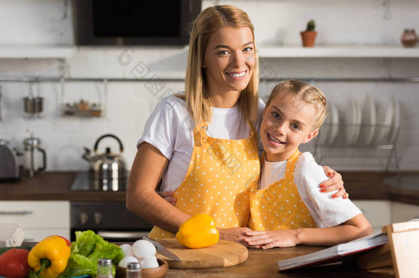 美丽快乐的母亲和女儿在围裙微笑着在相机, 而烹饪在一起 