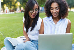 关闭两个欢快的女孩学生坐在草地上在校园里, 用笔记本电脑学习