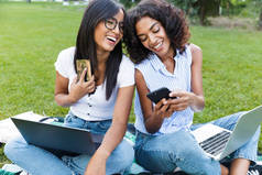 两个微笑的年轻女孩学生坐在草地上的校园, 学习, 手持手机
