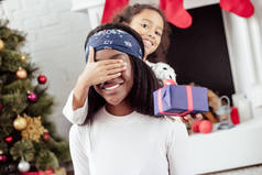 微笑的非洲裔美国女儿赠送圣诞礼物给母亲和关闭她的眼睛在家里惊讶