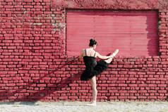 女芭蕾舞演员的背观, 在红砖墙前伸展, 她身穿黑色短裙。文本空间.