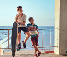 两个小兄弟摆在栅栏附近的一个海岸的背景上的滑板.
