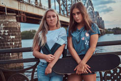 两个年轻的时髦女孩的肖像举行滑板和倚在旧桥背景上的护栏上.