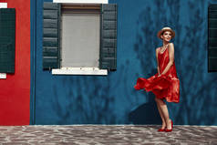 Fashoinable 穿着红色连衣裙和高跟鞋的年轻女子头戴草帽, 在布拉诺岛上摆出蓝色和红色的房子。墙上有树荫