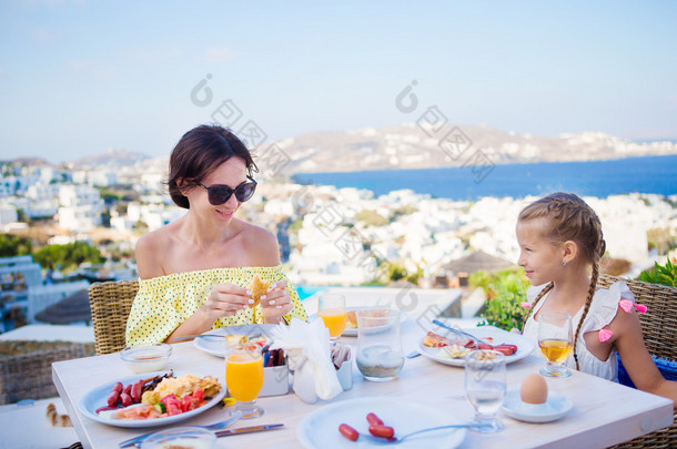 家庭在户外咖啡馆与风景米科诺斯小镇上吃早餐。可爱女孩和妈妈喝新鲜果汁，在豪华酒店的露台上吃牛角面包