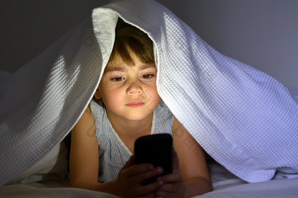 晚上，小孩在被子下面的床上玩智能手机