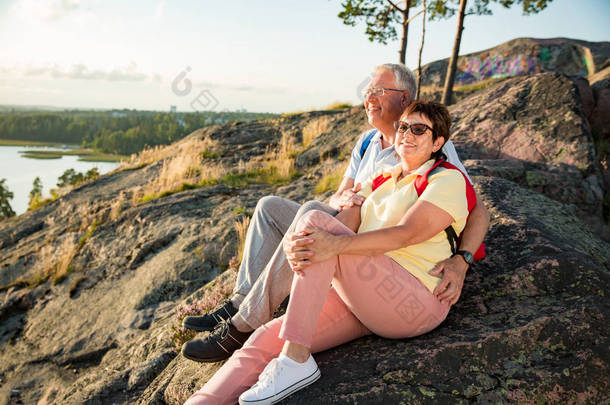 爱的老夫妇徒步, 坐在岩石的顶端, 探索。活跃成熟的男人和女人拥抱和愉快的微笑。海湾和海洋的风景。健康的生活方式。芬兰.