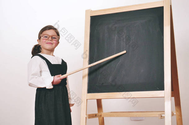 一个快乐的小女孩在一块小黑板前打扮成老师, 掌握着经济学、市场营销、团队合作、数学等课程。理念: 教育、学校、商业、热爱学习.