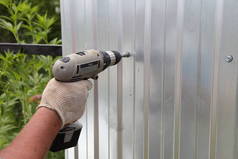 在阳光明媚的夏日，一个戴着工作手套的男人用电钻螺丝刀把螺丝拧在新的灰色金属板上，安全地建造花园篱笆