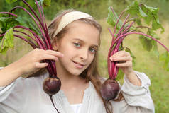 10岁的白人女孩双手捧着2根甜菜，背景是绿色植物