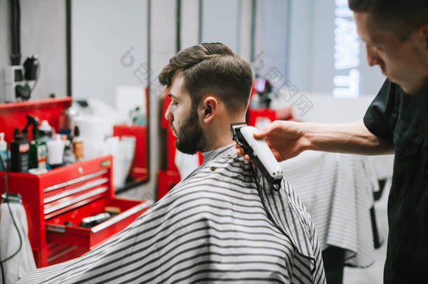 男式理发师为一个留着胡子的成年男子在浅色理发店里创造了一种时尚的发型。理发师用剪子剪断客户的头发.理发店概念.