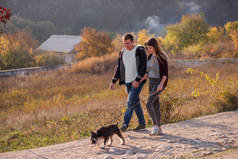 一个快乐的年轻家庭牵着一只雪纳犬沿着大路走着，手牵着手，在蓝天和森林的背景下彼此凝视着。有爱心的夫妻和宠物乘坐汽车周游世界