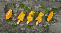 关闭三个水果冰激凌冰棒与新鲜橙片，绿色薄荷叶和冰块在灰色的桌子表面，高耸的顶部视野，直接上方