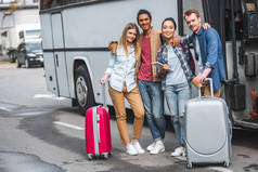年轻快乐多文化的朋友与轮袋摆在附近的旅游巴士在街道 