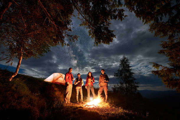朋友有休息的篝火和橙色帐篷在<strong>晚上</strong>附近的<strong>森林</strong>在多云的天空对远处山脉的剪影.