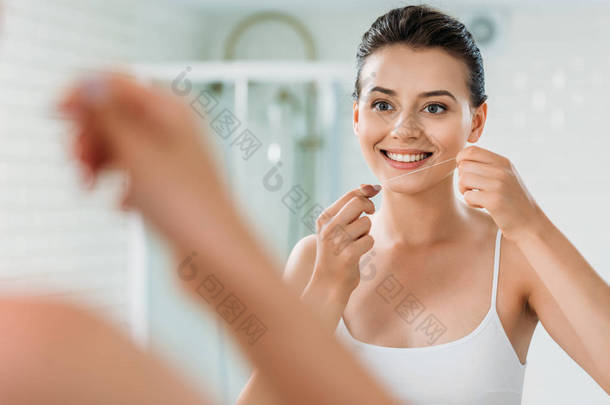 微笑的年轻妇女拿着牙线并且看镜子在洗手间 