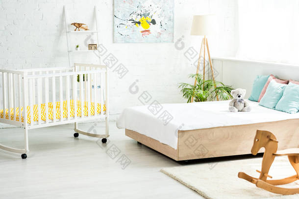 婴儿床靠近床与白色床上用品和枕头附近的泰迪熊和摇马 