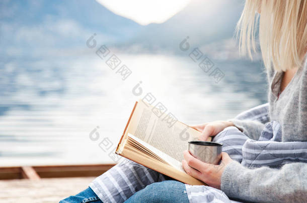 女孩正在海边的木<strong>码</strong>头上看书，冬天的大海，山，海滩。舒适的野餐，有热热的热蒸饮料，茶，咖啡或可可在热水瓶，温暖的格子。享受生活、放松的理念.