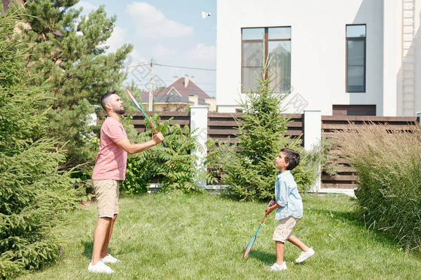 年轻的成年人穿着休闲装，和他的小儿子在后院草坪上打羽毛球，侧视拍摄