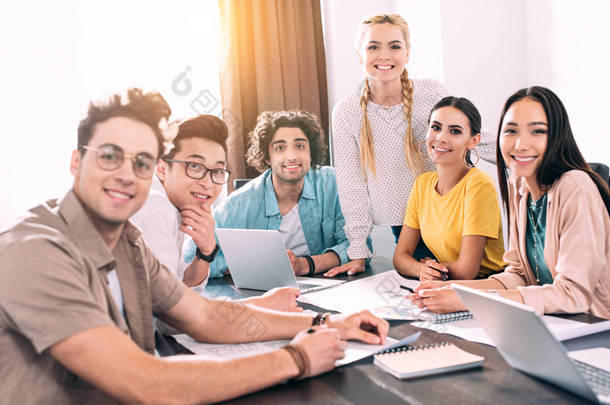 在现代办公室开会时，一群面带微笑的多文化商业伙伴与笔记本电脑和纸张坐在一起 