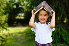 孩子女孩与书在她的头在户外。女生肖像, 教育理念。快乐的微笑可爱的孩子与书