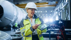专业的重工业女工程师身穿安全制服，手持数码台式机，并解释产品设计。石油、天然气和燃料输送管工业厂房建设