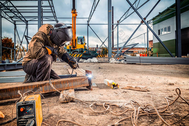 一个<strong>年轻男子</strong>焊工在棕色制服, 焊接面具和焊工皮革, 焊接金属与电弧焊机在建筑工地上, 蓝色火花飞向两侧