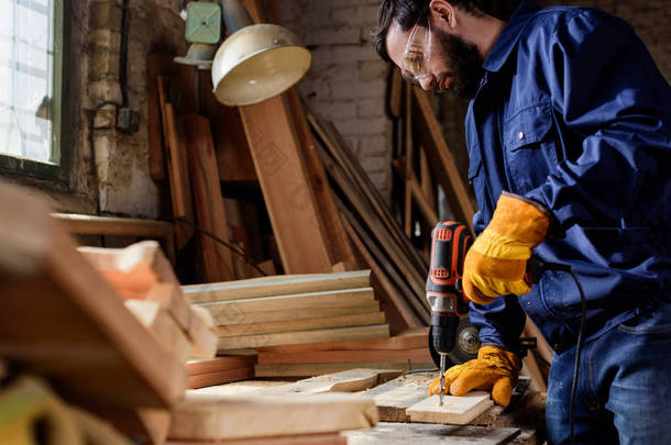 有胡子的工匠在防护手套和谷歌钻木板材在木工 