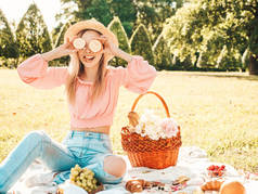 年轻美丽的嬉皮士女人穿着时髦的夏季牛仔裤，粉色T恤和帽子。外面野餐的女人正面的模特坐在草地上，吃着水果和奶酪，手里拿着柠檬 