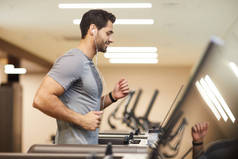 侧视图肖像英俊的年轻人在健身房的跑步机上跑步，微笑着享受锻炼，复制空间