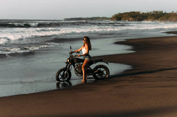美丽的单身女孩在海滩上骑摩托车与日落相遇。在海边骑自行车的时髦女人.骑摩托车旅行。去巴厘岛的旅行热带海滩上的一辆定制摩托车.复制空间
