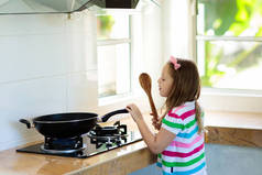 儿童烹饪。孩子们学会在炒锅里做蔬菜。小女孩用木勺搅拌油炸面条在锅里的煤气灶上。现代厨房内饰。儿童学习准备膳食