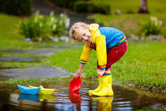 孩子们在水坑里玩纸船.孩子们在秋雨前在户外玩耍.为幼儿安排雨天户外活动。小孩在泥泞的水坑里跳跃。婴儿防水衣和靴子.
