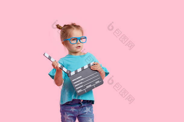 戴著电影眼镜的笑容可亲的小女孩拿着电影胶片,用粉红的背景把拍板隔开. 工作室肖像。 童年生活方式概念。 tex的复制空间
