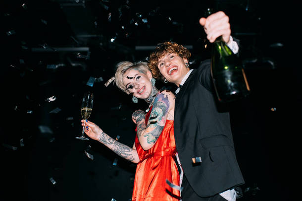 快乐的女人在红色礼服拿着一杯香槟酒在男朋友附近与瓶和微笑在黑色背景