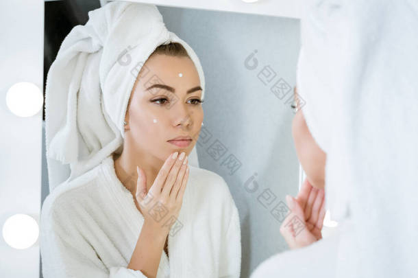 一位<strong>快乐</strong>的年轻女子在镜子前披着毛巾，在脸上涂上奶油，这在家里是个护肤的概念