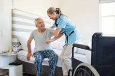 微笑的护士帮助老人起床从床上。护理护士支持病人, 而起床, 并向轮椅在家里。帮助老年残疾人站在他的卧室里.