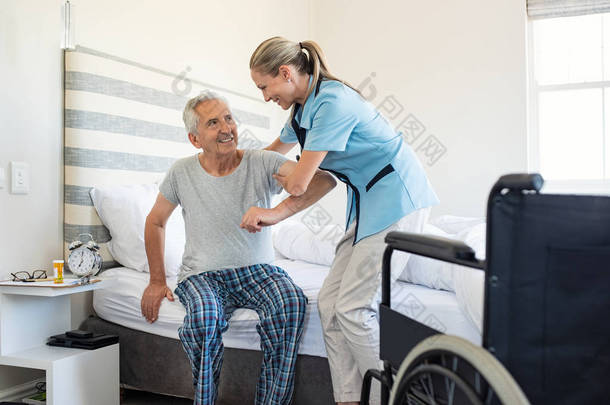 微笑的<strong>护士</strong>帮助老人起床从床上。<strong>护理护士</strong>支持病人, 而起床, 并向轮椅在家里。帮助老年残疾人站在他的卧室里.